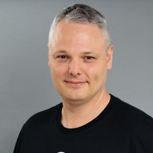 A middle aged white man with greying hair and a black tshirt against a grey background. 