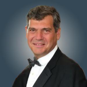 A middle aged white man with short salt and pepper hair at a ¾ turn in a black tuxedo and black bowtie. 