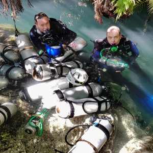 Two people in diving gear emerging from the water surrounded by oxygen tanks in an outdoor environment.