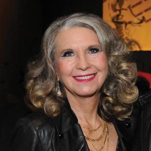 A middle aged white woman with curly grey hair smiles in a darkened room. 