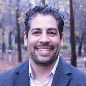 A hispanic man with facial hair wearing a wool suit jacket and collared shirt smiles outside in a wooded area 