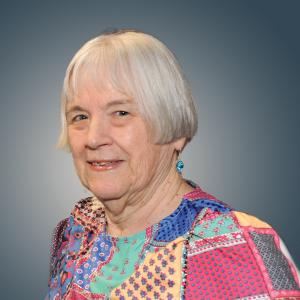 An elderly white woman with short cut to the ear white hair wearing a multicolor patchwork top. She is smiling and facing the camera at t ¾ turn. The background is a radial gradient of light to dark grey from center to outer.