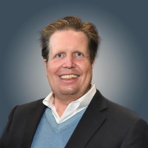 A middle aged white man in a grey suit jacket and light blue sweater with white color smiles at the camera.