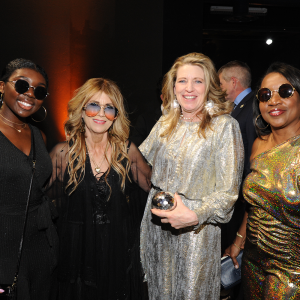 Two black women either side two white women all wearing 70s attire pose together and smile at the camera
