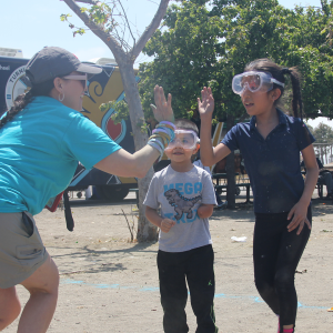 A young girl wearing safety goggles high-fives an adult woman while standing with a younger boy also wearing safety goggles