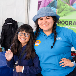 A young girl with bangs and dark hair and braces smiles shyly while a woman smiles with her arm around the girl