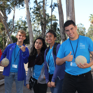 A group of teens, two boys and two girls smile at the camera outside. The boys are holding cantaloupes and flashing a shaka and thumbs up.