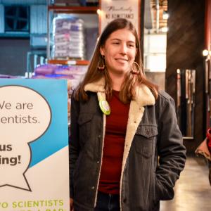 A woman in a sheepskin jacket stands near a sign that reads, "We are scientists. Ask us anything" at a brew pub. 