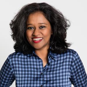 An indian woman with shoulder length dark hair wearing a blue checkered collared shirt smiles casually in front of a grey background 