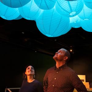 A woman and man in a darkened room stare up at a cluster of round blue lanterns 