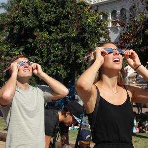 A man and woman wearing eclipse sunglasses squint up at the sun in a green outdoor area