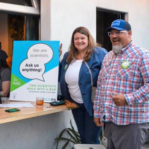 A woman and man stand outside a bar near a sign that says We are Scientists Ask Us Anything