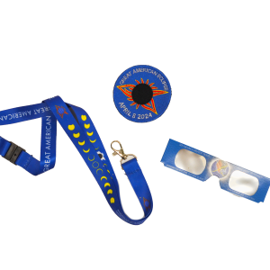 An aerial view of three items. A blue lanyard with a gold clip and a pattern of an eclipsing sun along the lanyard. A blue circle patch with a red arrowhead on the top and a black circle in the middle. And a pair of blue eclipse sunglasses. 