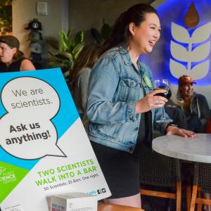 A sign in the foreground says We Are Scientists Ask Us Anything. A woman in the background holds a drink and talks with a smile