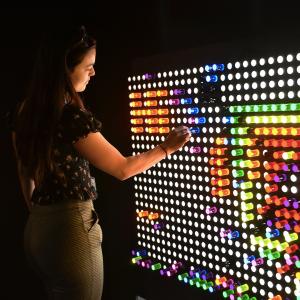 A woman standing in the dark playing with a huge lite brite on a wall