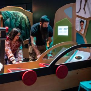 A girl and man interacting with a science exhibit shaped like a car