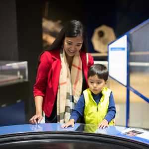 Mother wearing a red blazer and scarf and son wearing a yellow vest and a blue shirt play with an exhibit