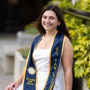 A white woman with dark hair wearing a white dress and a UC San Diego graduation stole outside 