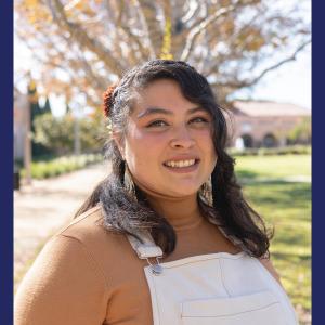 A hispanic woman in a brown top and white overalls outdoors in the fall smiling 