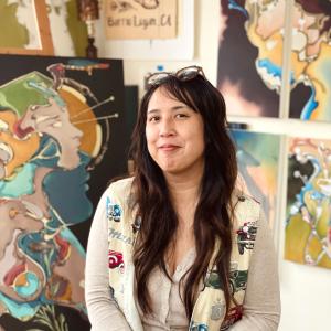 The photograph is a waist-up shot of a smiling woman with long, dark hair. The woman is looking directly at the camera. A pair of tortoiseshell glasses are perched atop her head. She is wearing a form-fitting, earth tone cardigan, slightly unbuttoned at the top. Over the cardigan, she sports a light-colored vest decorated with various vintage car illustrations.  Behind her, the space transitions into an artistic studio or gallery. The backdrop is adorned with numerous abstract and illustrative paintings. To
