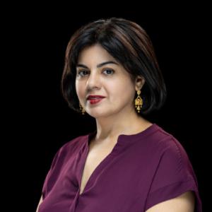 A medium-shot studio portrait of a light-skinned, middle-aged woman with straight, chin-length black hair parted to her left, wearing a deep plum-colored, loose-fitting blouse, and gold, ornate dangling earrings set against a solid black background.