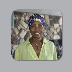 A portrait of a smiling Black woman with an oval face wearing a patterned headwrap, a collared blouse, and a necklace dominates a square-format photograph set against a soft gray background. The woman's headwrap features a royal blue background adorned with large, light green, amoeba-like shapes. She is wearing a lime green button-down blouse, its collar open just enough to reveal a hint of a pink lining. A delicate necklace encircles her neck, featuring small silver beads alternating with larger, darker on