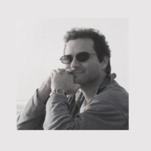 A black and white photo shows a light-skinned male sitting with his knees up while wearing sunglasses. The subject has dark curly hair that is close to his head with some loose curls on his forehead. He is wearing dark sunglasses with circular rims. His hands are clasped together and positioned near his mouth. His right wrist has a wristwatch with a light-colored band. He's wearing a long-sleeved shirt or light jacket over a dark undershirt. The shirt is buttoned up to the middle of his chest. He is wearing