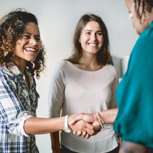 In a brightly lit gathering, three women are engaged in a warm and professional interaction. The focal point is the handshake between a woman on the left, dressed in a vibrant white and blue plaid shirt, and a woman on the right, partially visible with a striking teal blouse. The woman on the left has curly, dark hair with brown highlights, and a radiant smile that reveals her teeth. She is wearing a white watch with a rose gold face, adding a touch of elegance to her casual attire.