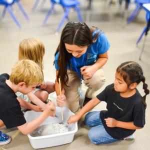 In a bright indoor setting, a fair-skinned, young adult woman with long, dark brown hair and three young children participate in a science project, gathered on the floor around a white plastic container. The woman, likely a science volunteer, assists the children with the experiment, holding a slender wooden rod, possibly a stirring stick, and peering down into the container. She is dressed in a blue short-sleeve shirt featuring a science-related logo and the words "SCIENCE VOLUNTEER," paired with tan khaki