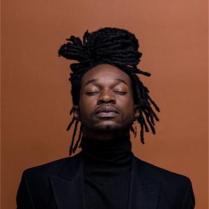 A man with his eyes closed and dreadlocks wearing a black suit stands confidently, showcasing a professional appearance.
