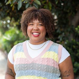 A woman with curly hair is dressed in a bright, multicolored sweater, exuding a cheerful demeanor.